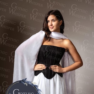 Op de afbeelding: Een vrouw draagt een zwart korset, een witte rok en een witte cape. Het korset heeft een gestructureerd ontwerp en de rok is vloeiend. De cape is over de schouders gedrapeerd. De achtergrond heeft een herhalend patroon van de woorden "Gothic Whisper".