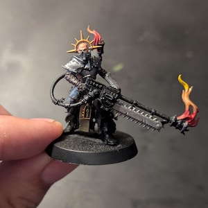 May include: A painted miniature figurine of a warrior wearing dark armour and a gold halo-style headpiece. The figure holds a long black weapon with a chainsaw blade and a flame effect at the tip, with an additional flame rising from the back.