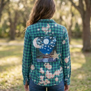 May include: A green and navy blue plaid long-sleeve button-down shirt with a bleached pattern. The back features a rectangular patch depicting a night landscape with a moon, stars, a mountain range, a lake, and a large yellow star overlay.