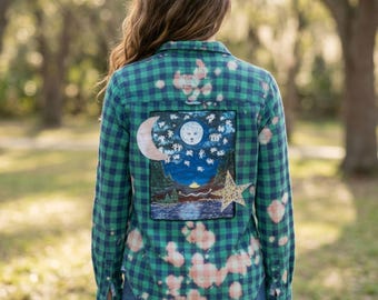 May include: A green and navy blue plaid long-sleeve button-down shirt with a bleached pattern. The back features a rectangular patch depicting a night landscape with a moon, stars, a mountain range, a lake, and a large yellow star overlay.