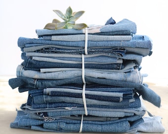 May include: A stack of assorted blue denim fabric scraps and pieces of varying shades, textures, and washes, neatly bundled together with a white elastic cord. A small, light green succulent plant rests on the very top of the denim stack.