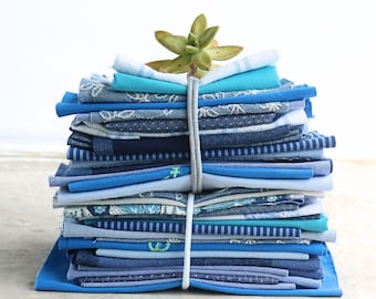 May include: A stack of folded fabric squares in various shades of blue, including navy, sky blue, and turquoise. The fabrics feature diverse patterns such as stripes, floral prints, and solid colors, all secured together by a light gray elastic band.