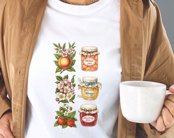 May include: White t-shirt featuring watercolor illustrations of peach, honey, and strawberry jam jars. Each jar is labeled and surrounded by floral and fruit elements. The shirt is worn by a person holding a white mug.