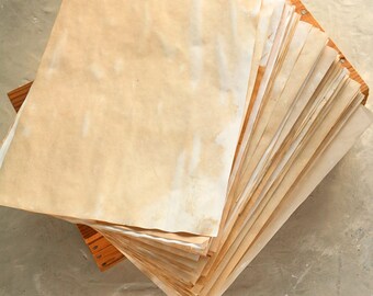 May include: A stack of aged, stained paper sheets, possibly parchment or vintage paper, arranged on a wooden base. The paper has a warm, yellowish-brown hue with darker spots, suggesting age or staining. The sheets are stacked at an angle.
