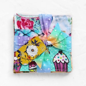 May include: A stack of fabric coasters tied with a rainbow polka dot ribbon and a yellow floral tag. The coasters feature a variety of colorful patterns, including roses, cupcakes, and polka dots. The tag has a white flower design.