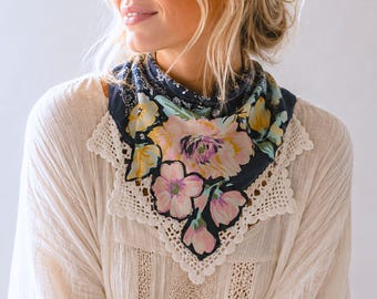 May include: A triangular bandana featuring a dark navy blue background with a vibrant floral print in shades of pink, yellow, and green. The edges of the bandana are finished with a wide, cream-colored crocheted lace trim with a scalloped border.