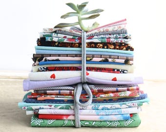 May include: A stack of assorted fabric swatches in various colors and patterns, including floral, heart, and plaid prints, tied together with a grey fabric strip. A small green succulent plant sits on top of the folded textile bundle.