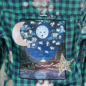 May include: Green and blue plaid flannel shirt with a painted patch featuring a wolf in a moon, surrounded by stars and forest scenery. A crescent moon and a floral star are also attached to the shirt.
