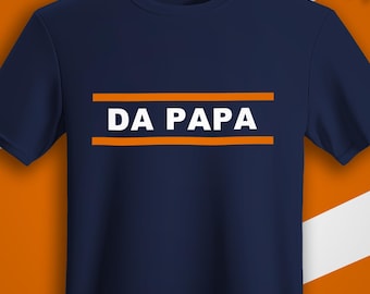 May include: Navy blue t-shirt with the text "DA PAPA" in white letters, framed by two orange lines. The shirt is hanging on a wooden hanger against an orange background. The shirt has a round neck and short sleeves.