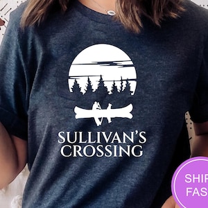 May include: Dark blue t-shirt featuring a white graphic of a canoe with two people, trees, and a sun. The text "SULLIVAN'S CROSSING" is below the graphic. The shirt has short sleeves.
