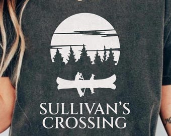 May include: Dark gray t-shirt featuring a white graphic of a canoe with two people, trees, and a sun. The text "SULLIVAN'S CROSSING" is printed below the graphic in a clean, sans-serif font. The shirt has a crew neck and short sleeves.