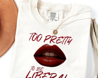May include: Off-white t-shirt with the text "TOO PRETTY TO BE LIBERAL" in red. The shirt features a graphic of red lips. A gold necklace and a brown hat are visible in the image.
