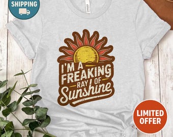 May include: A light gray t-shirt with a graphic that reads "I'M A FREAKING RAY OF SUNSHINE." The graphic features a sun design in shades of brown, yellow, and red. The shirt is a casual style.