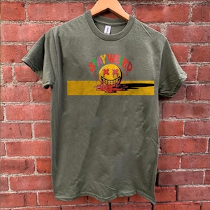 Può includere: Una maglietta verde con una grafica gialla, rossa e verde di un viso sorridente che si scioglie con il testo "STAY WEIRD" sopra.