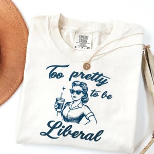 Può includere: T-shirt color crema con la scritta "Too pretty to be Liberal" in blu scuro. La grafica presenta una donna in stile retrò con occhiali da sole che tiene in mano una bevanda. Una collana d'oro è appoggiata sulla maglietta.