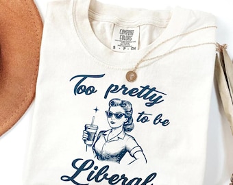 May include: Cream-colored t-shirt with the text "Too pretty to be Liberal" in dark blue script. The graphic features a retro-style woman in sunglasses holding a beverage. A gold necklace rests on the shirt.