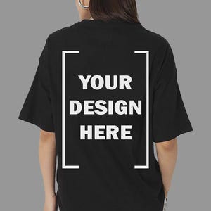Può includere: T-shirt nero con la scritta "YOUR DESIGN HERE" in un riquadro rettangolare bianco. La maglietta ha maniche corte e una vestibilità ampia. La persona indossa orecchini a cerchio argentati.