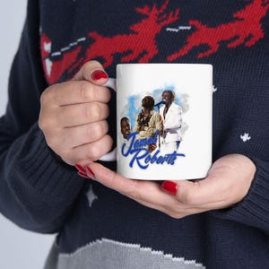 Può includere: Tazza in ceramica bianca con una grafica di tre persone e la scritta "Jamal Roberts" in blu. La tazza è tenuta da una persona che indossa un maglione blu con un motivo a renne.
