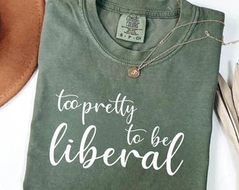 May include: Olive green t-shirt with the text "too pretty to be liberal" in white script. A silver necklace with a small pendant is draped on the shirt. The shirt is folded and displayed on a white surface.