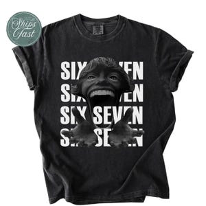 Può includere: T-shirt nera con un design grafico raffigurante un volto distorto e le parole "SIX SEVEN" a blocchi bianchi. La maglietta ha maniche corte e girocollo.