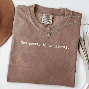 Può includere: Una maglietta marrone Comfort Colors con la scritta "Too pretty to be liberal." Una collana d'argento con un ciondolo è drappeggiata sulla maglietta piegata. La maglietta ha un'etichetta sul collo.