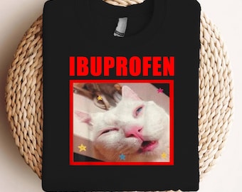 May include: Black t-shirt featuring the word "IBUPROFEN" in red capital letters. Below is a photo of a white cat with its eyes closed and mouth slightly open, framed in red, with star graphics.