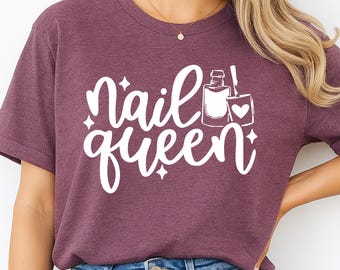 May include: A maroon t-shirt with the words "Nail Queen" in a white, cursive font. The design includes a nail polish bottle, a nail polish brush, and a heart. The shirt is made of a soft material.