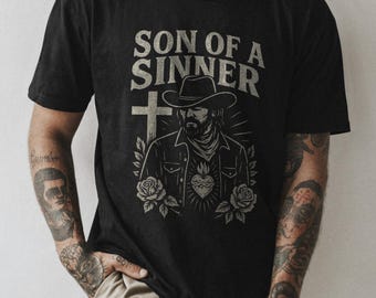May include: Black t-shirt with a graphic print of a man wearing a cowboy hat and a cross. The text "Son of a Sinner" is printed above the graphic.