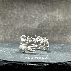 May include: A silver ring with a detailed, organic design, possibly representing foliage. The ring is presented on a dark, textured surface. The text "LENA WONG Art Jewelry Design" is visible.