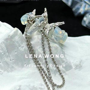 May include: A silver-toned earring with a starburst design and dangling ball chain. The earring features a cluster of opalescent stones and the text "LENA WONG Art Jewelry Design".
