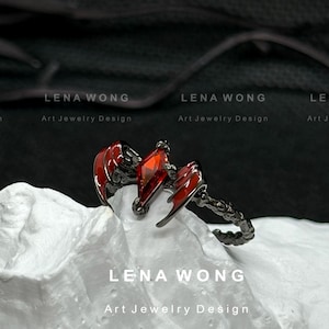 Może przedstawiać: Czarny pierścionek z czerwoną szlachetnym kamieniem i akcentami w kształcie skrzydeł. Pierścionek jest prezentowany na białej powierzchni. Widoczny jest tekst "LENA WONG" i "Art Jewelry Design".