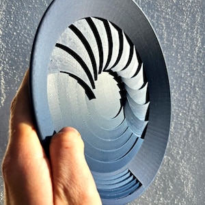 May include: A circular dark gray object featuring a mechanical iris design with multiple overlapping curved blades that form a central aperture. The structure is constructed from layered segments that create a radial pattern.