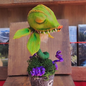 May include: A whimsical, handmade sculpture of a green plant-like creature with a toothy mouth and closed eyes. The sculpture has green leaves, a green stem, and purple tendrils, set in a pot with green moss. The background includes wooden planks and postcards.