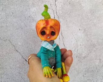 May include: A handmade Halloween-themed doll with a pumpkin head, large black eyes, and a small mouth. The doll wears a teal jacket, a blue and white checkered collar, and has green hands and feet. The pumpkin head is orange and green.