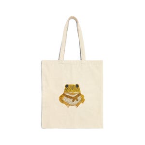 May include: A cream-coloured tote bag with a detailed illustration of a yellow and brown frog wearing a scarf. The frog is centered on the bag, with the handles extending upwards. The bag is made of a sturdy fabric.
