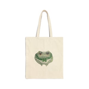 May include: Cream-colored tote bag featuring a detailed illustration of a frog wearing a green scarf. The frog has large, expressive eyes and a textured appearance. The bag has two long handles for carrying.