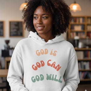 Könnte beinhalten: Ein weißer Kapuzenpullover mit dem Text "GOD IS GOD CAN GOD WILL" in Orange, Rot und Grün. Der Pullover hat eine Kapuze mit Kordelzug und eine Vordertasche. Die Person auf dem Bild lächelt.