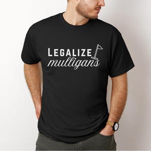 Peut inclure: T-shirt noir avec le texte blanc "LEGALIZEmulligans" et un graphique de drapeau de golf. Le t-shirt est à col rond et semble être en coton. Le texte est en cursive.