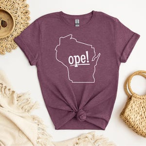 May include: A maroon t-shirt featuring a white outline of the state of Wisconsin with the word "ope!" in white text. The shirt is knotted at the bottom and is surrounded by decorative items.