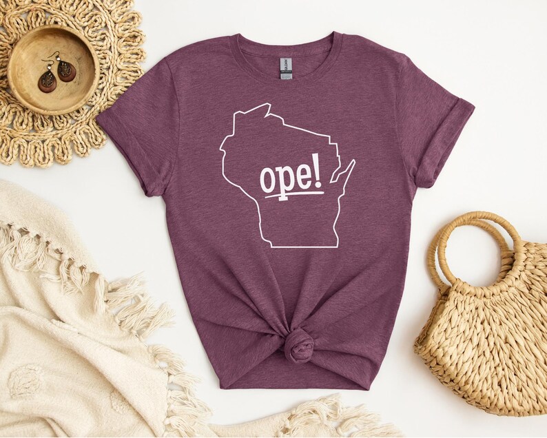 May include: A maroon t-shirt featuring a white outline of the state of Wisconsin with the word "ope!" in white text. The shirt is knotted at the bottom and is surrounded by decorative items.