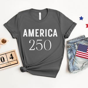 May include: Dark gray t-shirt with the text "AMERICA 250" in white. The shirt is styled with denim shorts, an American flag, wooden sunglasses, and a wooden calendar set to July 4th. Red and blue star decorations are also present.