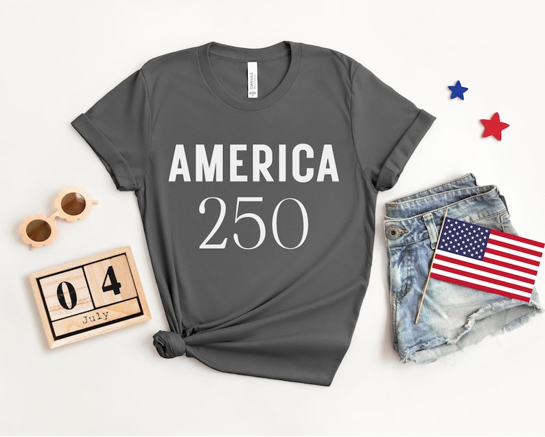 May include: Dark gray t-shirt with the text "AMERICA 250" in white. The shirt is styled with denim shorts, an American flag, wooden sunglasses, and a wooden calendar set to July 4th. Red and blue star decorations are also present.