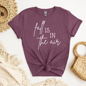 May include: A maroon t-shirt with the text "Fall is in the air" in white script. The shirt is knotted at the waist. A woven straw bag and decorative items are in the background.