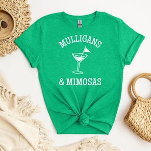 May include: A green t-shirt with the words "Mulligans & Mimosas" in white, along with a cocktail glass graphic. The shirt is knotted at the waist. A straw bag and decorative items are in the background.