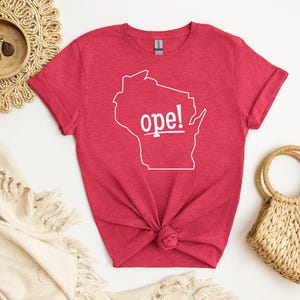 May include: A red t-shirt with a white outline of the state of Wisconsin and the word "ope!" in white. The shirt is knotted at the bottom and is surrounded by a woven placemat, a small wooden bowl, and a straw handbag.