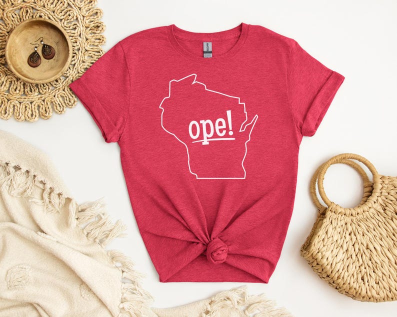May include: A red t-shirt with a white outline of the state of Wisconsin and the word "ope!" in white. The shirt is knotted at the bottom and is surrounded by a woven placemat, a small wooden bowl, and a straw handbag.