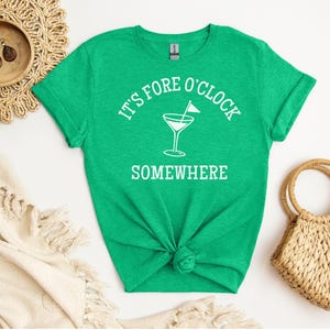 May include: A bright green t-shirt with the text "It's Fore O'Clock Somewhere" in white, along with a martini glass graphic. The shirt is knotted at the waist. A woven straw bag and decorative items are in the background.