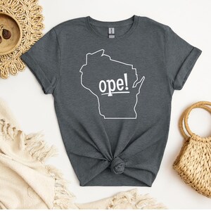 May include: Dark gray t-shirt featuring a white outline of the state of Wisconsin with the word "ope!" printed in white. The shirt is knotted at the waist and is displayed with a woven bag and decorative items.