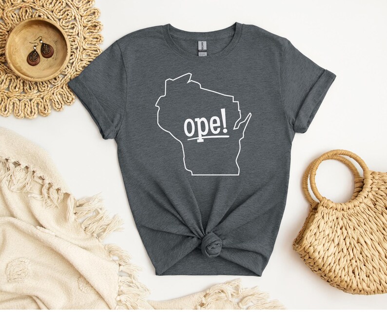 May include: Dark gray t-shirt featuring a white outline of the state of Wisconsin with the word "ope!" printed in white. The shirt is knotted at the waist and is displayed with a woven bag and decorative items.