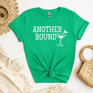 May include: A green t-shirt with the text "ANOTHER ROUND" in white, accompanied by a martini glass graphic. The shirt is knotted at the bottom. A woven straw bag and decorative plate are in the background.